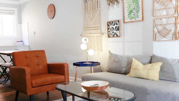 A modern living room with a sofa, armchair, coffee table, wall art, and a clock. - Sojourn Cove Suite 2 BR 2 BA at Beach Cottages (Norfolk)
