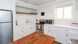 A modern kitchen with white cabinetry, a stainless steel refrigerator, a microwave, and a wooden dining table. - Sojourn Cove Suite 2 BR 2 BA at Beach Cottages (Norfolk)