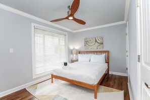 Une chambre avec un lit en bois, un ventilateur de plafond, une fenêtre avec des stores, une lampe de chevet et une œuvre d’art murale.