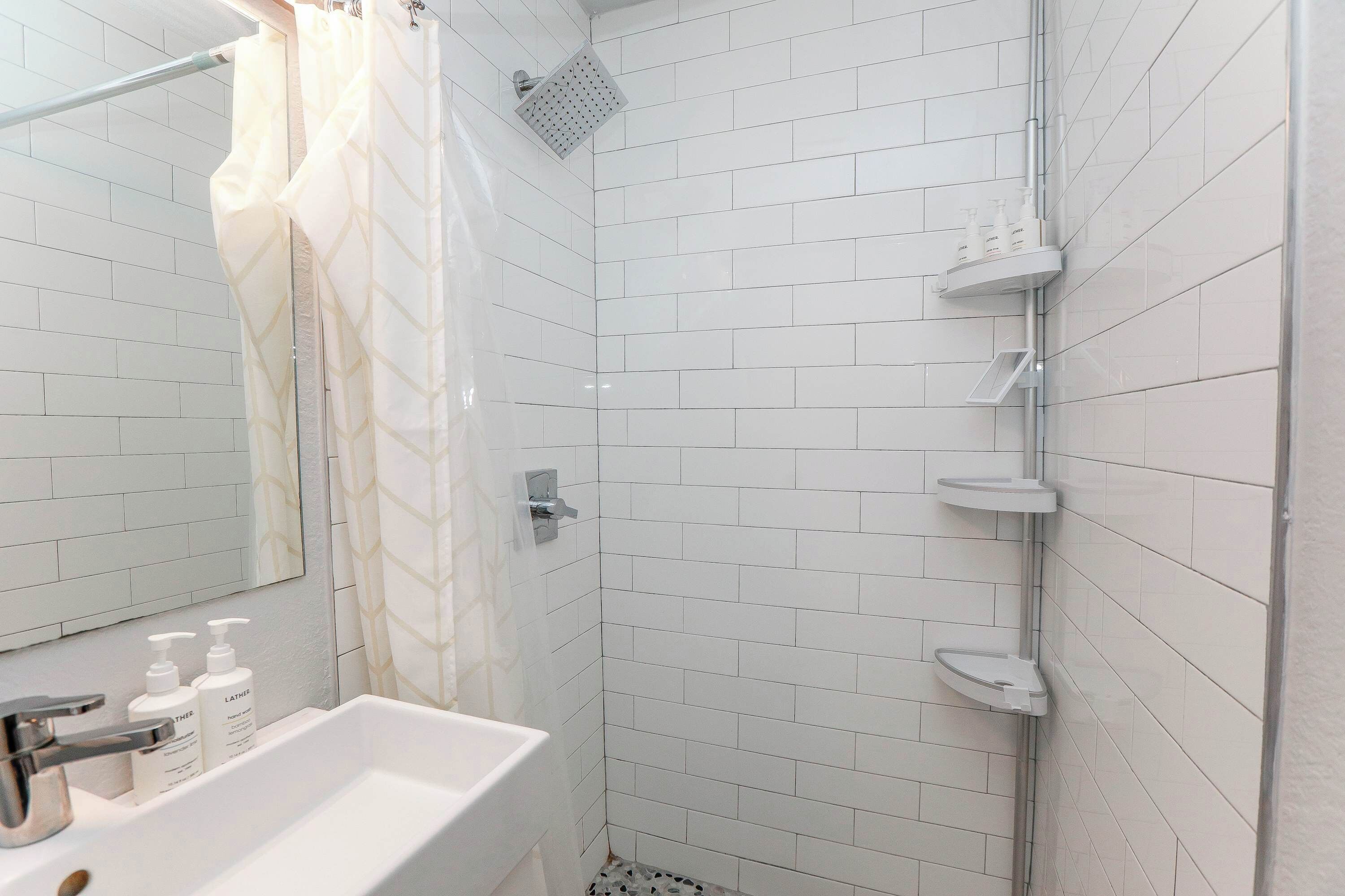 Un baño con paredes de azulejos blancos, una ducha con puerta de vidrio, un lavamanos con dos botellas de loción y un espejo.