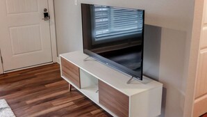 A modern living room with a flat-screen TV on a white TV stand, wooden flooring, and a white door. - Sojourn's Tide Suite at Beach Cottages (Norfolk)