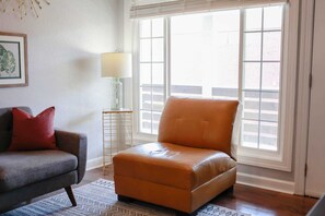 Ein Zimmer mit Ledersessel, grauem Sofa mit roten Kissen, Stehlampe und einem gemusterten Teppich.