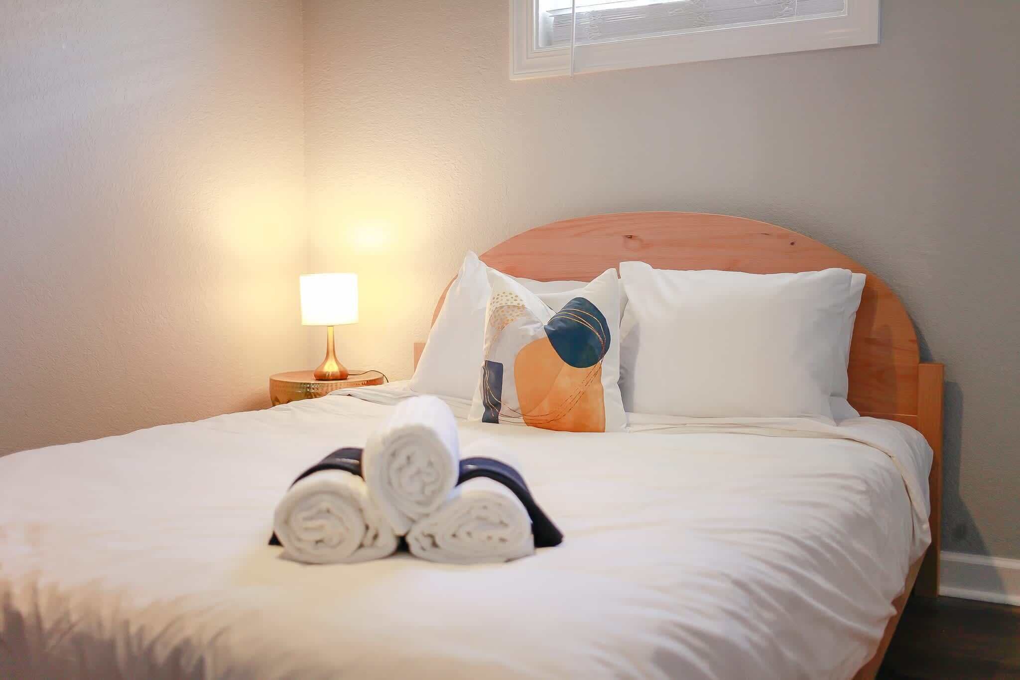 A neatly made bed with rolled towels, a wooden headboard, and a bedside lamp.