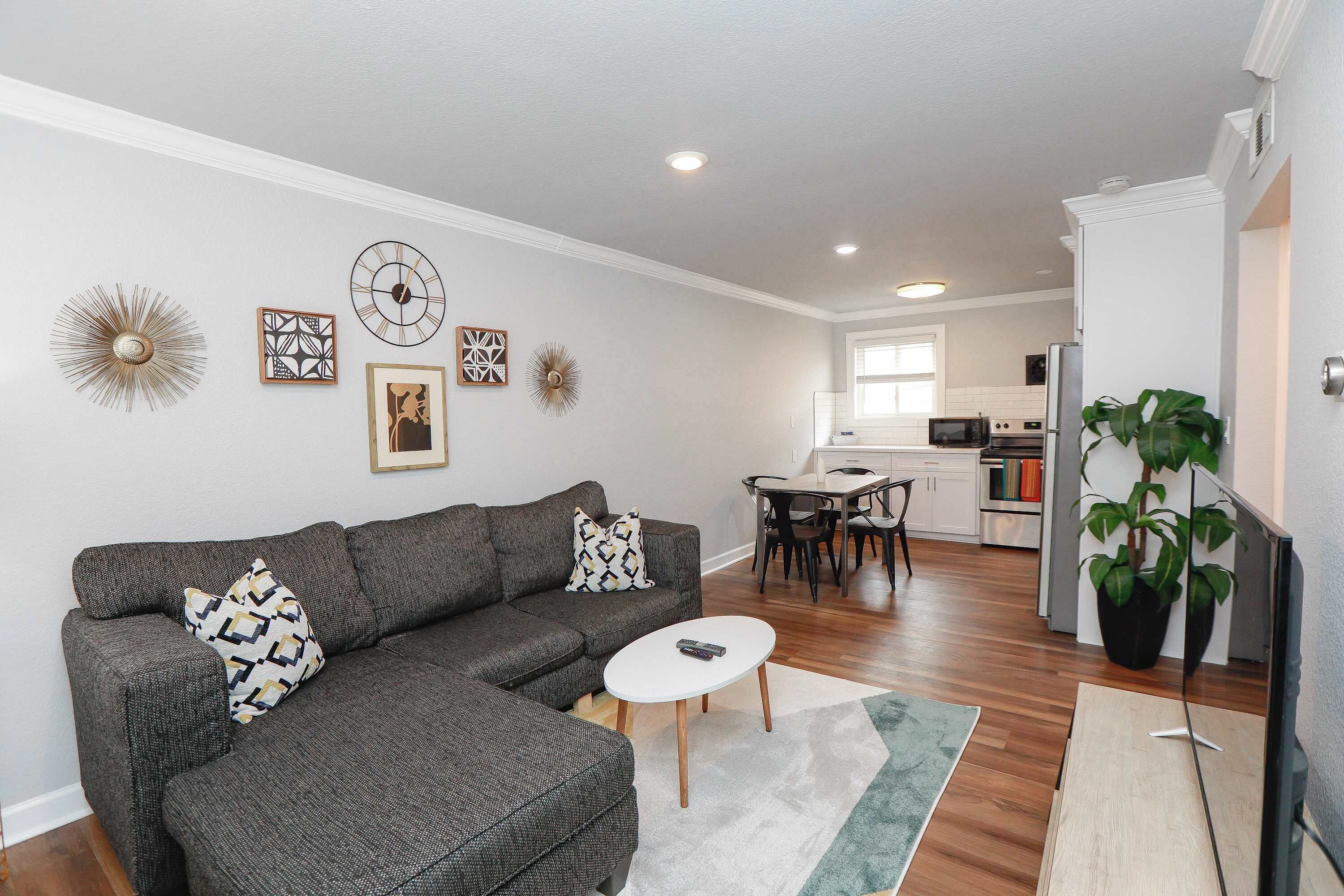 Un salon comprenant un canapé, une table basse et des éléments décoratifs sur les murs. On y trouve également un coin repas avec une table et des chaises, et une cuisine à l’arrière-plan.