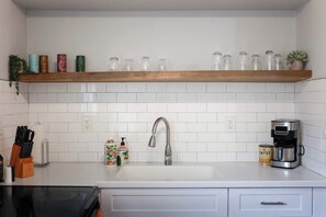 A kitchen with a white tiled backsplash, a stainless steel faucet, a coffee maker, and a shelf with glass jars. - Sojourn Tide Suite 2 BR 2 BA at Beach Cottages #11 (Norfolk)