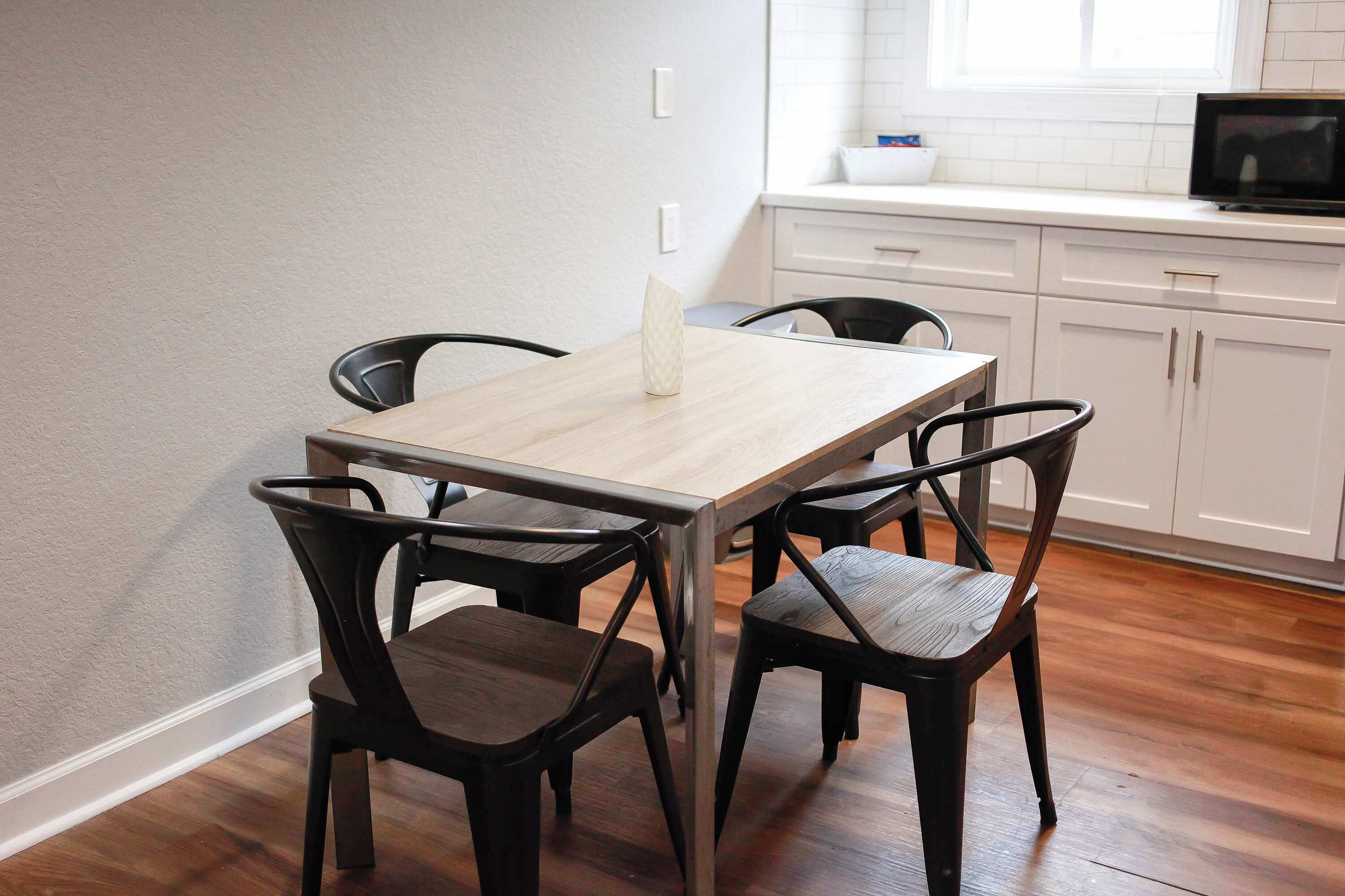A dining area with a wooden table and four chairs, a white countertop with a microwave, and a window with blinds.