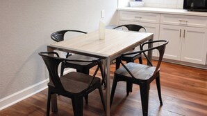 En matplats med ett träbord och fyra stolar, en vit bänkskiva med en mikrovågsugn och ett fönster med persienner.