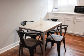 A dining area with a wooden table and four chairs, a white countertop with a microwave, and a window with blinds. - Sojourn Tide Suite 2 BR 2 BA at Beach Cottages #11 (Norfolk)
