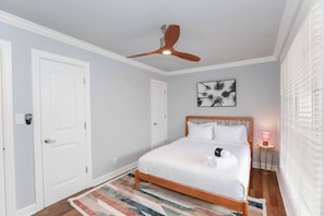 A bedroom with a wooden bed, white walls, a ceiling fan, a door, a framed picture, a bedside lamp, and a striped rug. - Sojourn Tide Suite 2 BR 2 BA at Beach Cottages #11 (Norfolk)