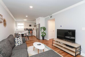 A modern living room with a sofa, coffee table, TV, and a dining area with a table and chairs. - Sojourn Tide Suite 2 BR 2 BA at Beach Cottages #11 (Norfolk)