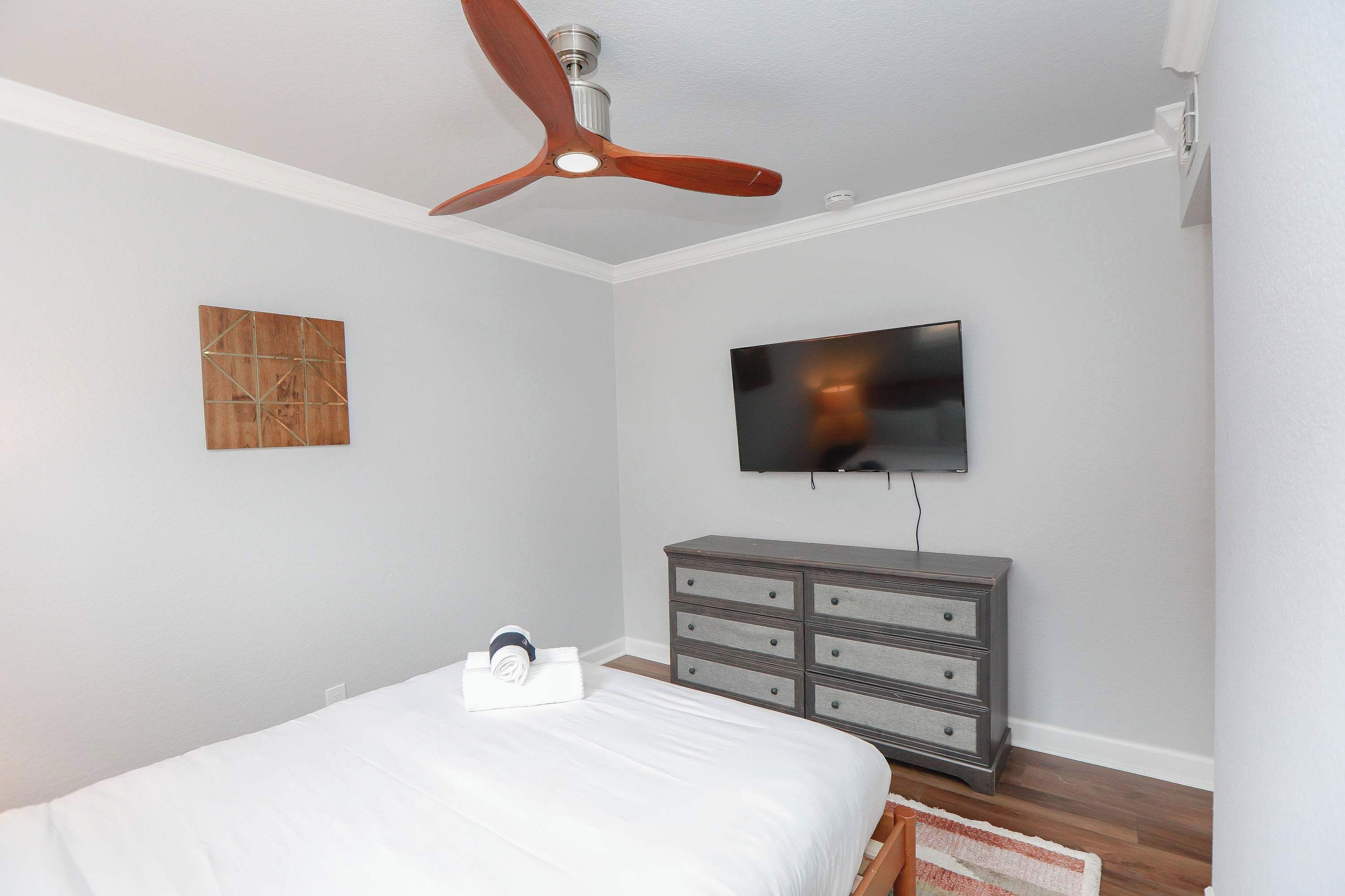 Une chambre à coucher comprenant un lit, une commode, un ventilateur de plafond, un téléviseur fixé au mur et une œuvre d’art décorative.