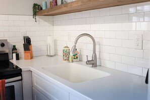 A kitchen with a white countertop, a stainless steel sink, and a wooden shelf above. - Sojourn Tide Suite 2 BR 2 BA at Beach Cottages #11 (Norfolk)