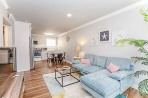 A modern living room with a blue sofa, a glass coffee table, and a dining area with a table and chairs. - Sojourn Tide Suite 2 BR 2 BA at Beach Cottages (Norfolk)