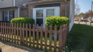A two-story house with a brick exterior, a wooden picket fence, and green shrubs. - Sojourn | Virginia Beach | 2 Bedroom Townhouse (Virginia Beach)