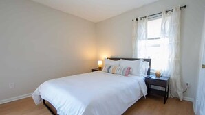 A bedroom with a single bed, a bedside table with a lamp, and a window with curtains. - Sojourn | Virginia Beach | 2 Bedroom Townhouse (Virginia Beach)