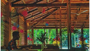 A restaurant with a wooden ceiling and large windows. There are wicker chairs and tables, and a person is seated at a table. - Angel Eco Resort & Restaurant Cox'sBazar (Cox's Bazar)