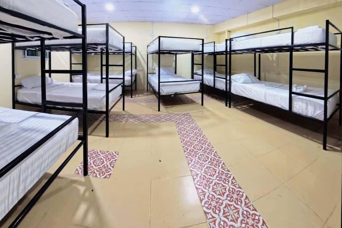A dormitory room with bunk beds arranged in two rows, a patterned carpet, and a tiled floor.