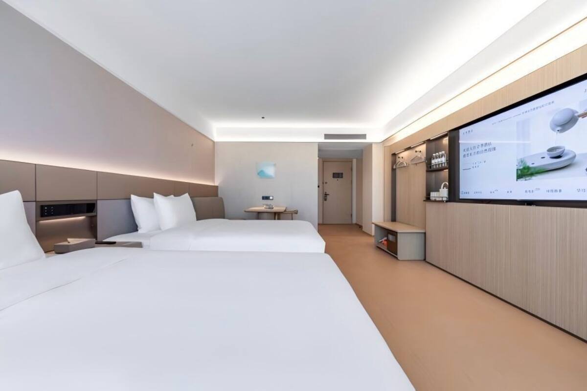 A modern hotel room with two beds, a small table, and a TV screen displaying a menu.