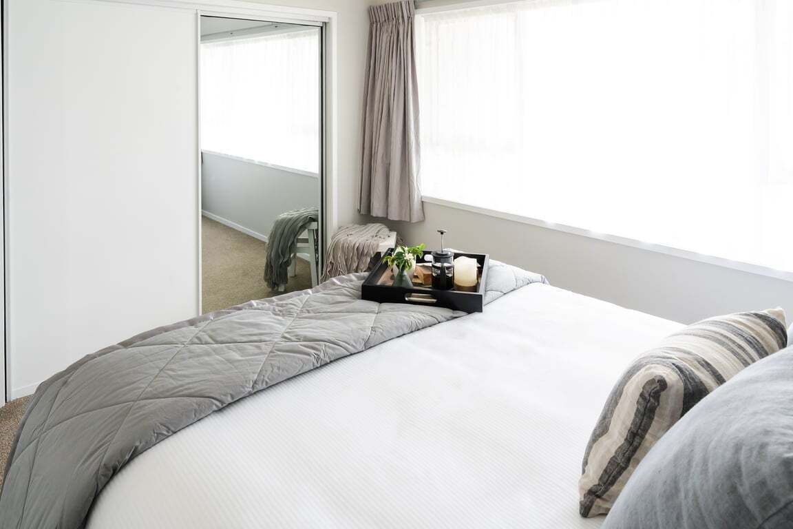 Una cama bien hecha con colcha gris, una mesita de noche con una bandeja de objetos y un espejo grande que refleja una habitación con ventana y cortinas.
