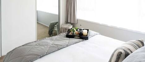 Una cama bien hecha con colcha gris, una mesita de noche con una bandeja de objetos y un espejo grande que refleja una habitación con ventana y cortinas.