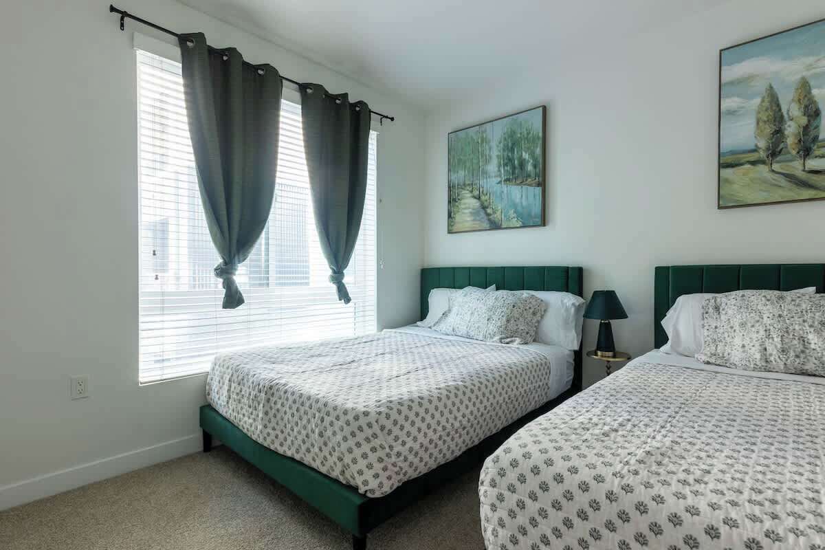 A bedroom with two beds, a window with curtains, a painting on the wall, and a lamp on a bedside table.