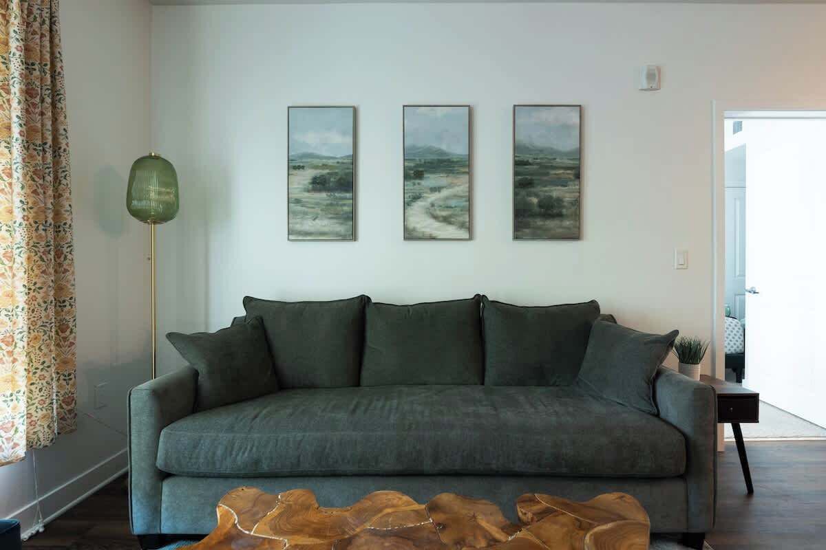 A living room with a grey sofa, three framed landscape paintings on the wall, a green floor lamp, and a wooden coffee table.