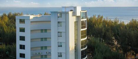 Un edificio de varios pisos con balcones, vista despejada al mar y zona boscosa al fondo.