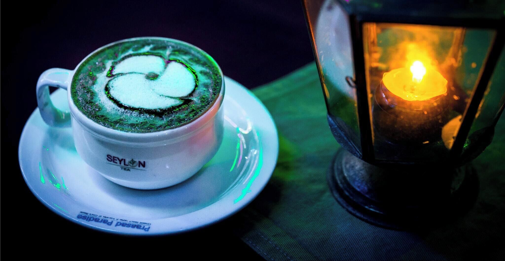 Une tasse de thé ornée d’un motif vert sur la soucoupe, une bougie dans une lanterne et un fond sombre.