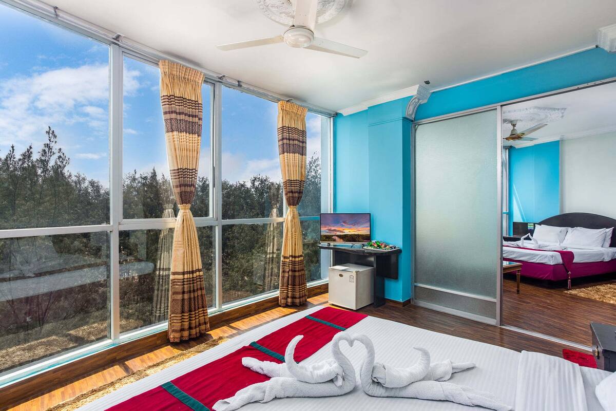 Una habitación de hotel moderna con un ventanal grande, una cama y un tapete rojo y blanco a rayas, un televisor y una puerta corredera que da a otra habitación.