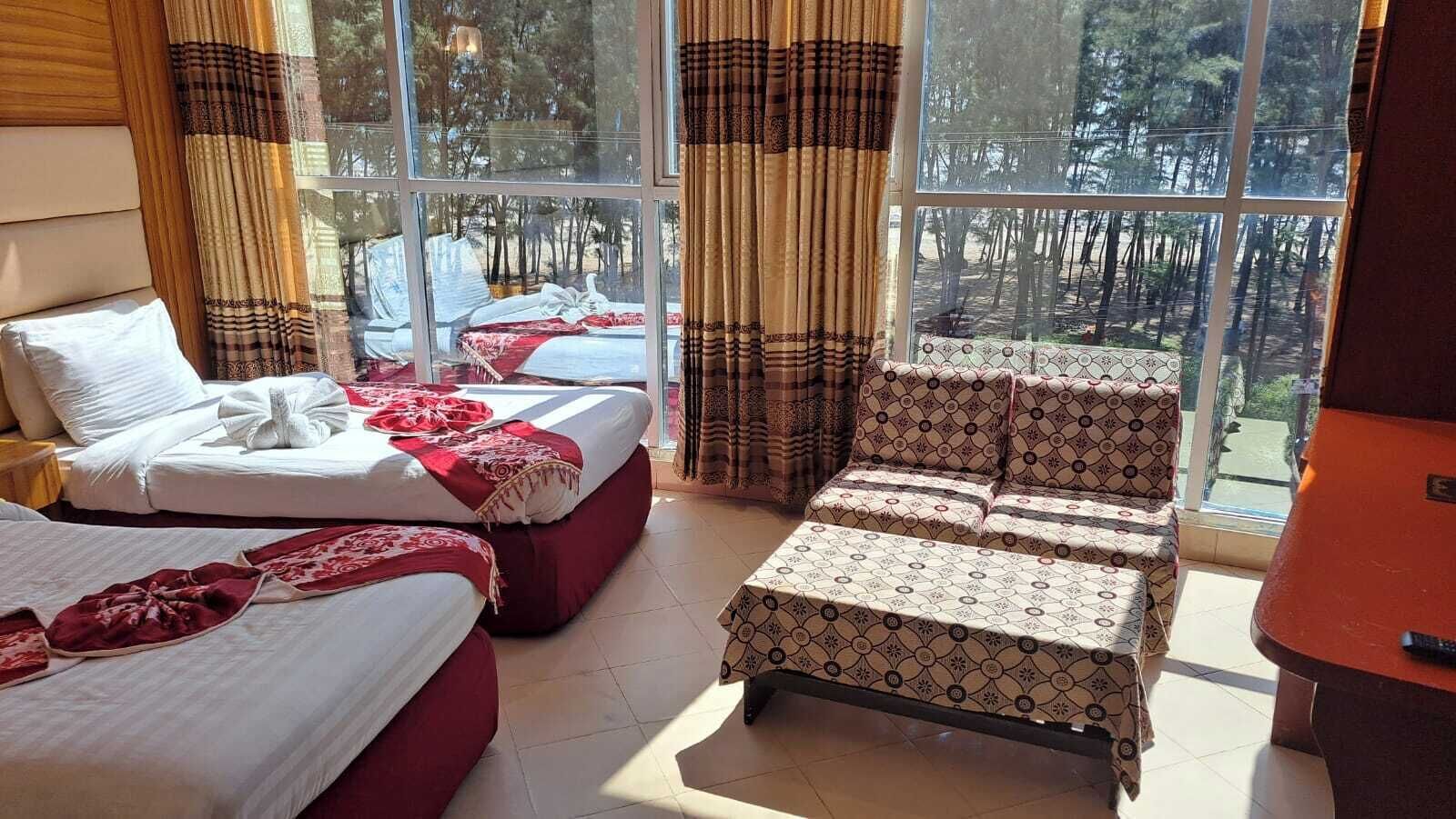 A hotel room with two beds, a sofa, and a large window with a view of trees.