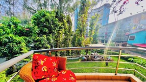 Ein Balkon mit Korbmöbeln, gefliestem Boden und Blick auf einen Garten mit Bäumen und einem Gebäude im Hintergrund.