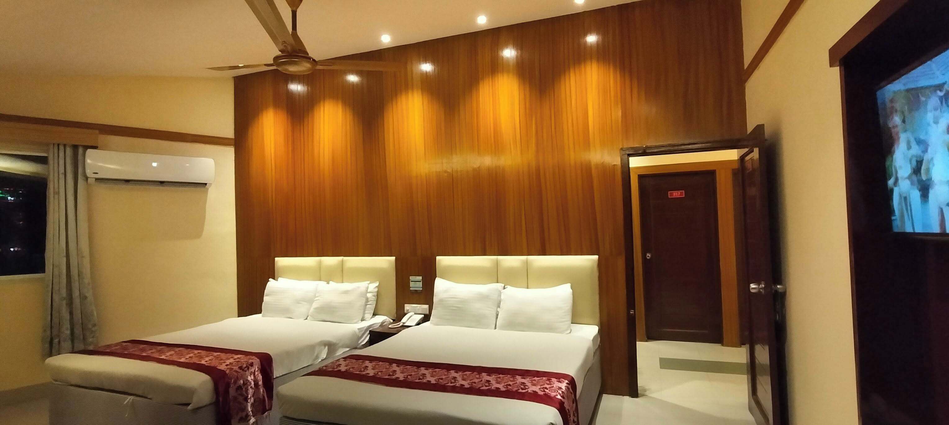 Una habitació d'hotel amb dos llits, un ventilador de sostre, un aparell d'aire condicionat, una televisió i un panell de paret de fusta.