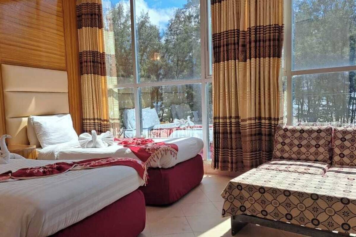 Habitación de hotel con dos camas, un sofá, un ventanal grande con cortinas y vistas a árboles.