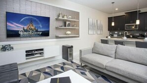 Una sala moderna con televisor de pantalla plana, sofá gris y área de cocina con armarios oscuros e iluminación colgante.