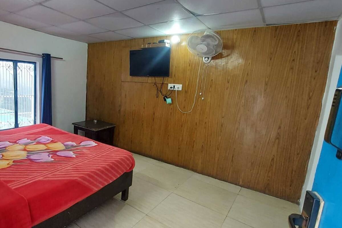 Une chambre avec un lit, un téléviseur fixé au mur, un mur recouvert de panneaux en bois, une fenêtre avec des rideaux et un ventilateur.
