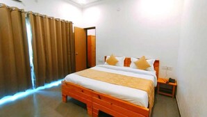 Un dormitorio con cama de madera, ropa de cama blanca y amarilla, una mesita de noche con teléfono y una cortina entreabierta.