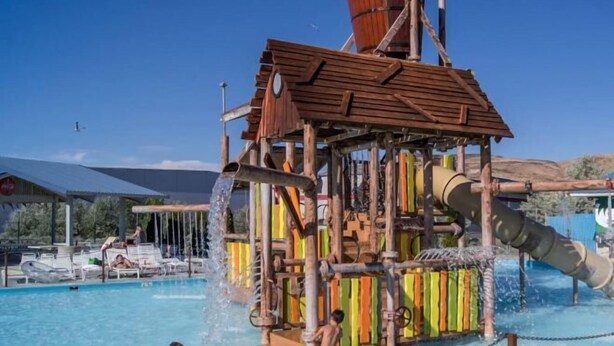 Una estructura de juegos acuáticos con techo de madera y paneles de colores, rodeada de una zona de piscina con sillones de descanso.