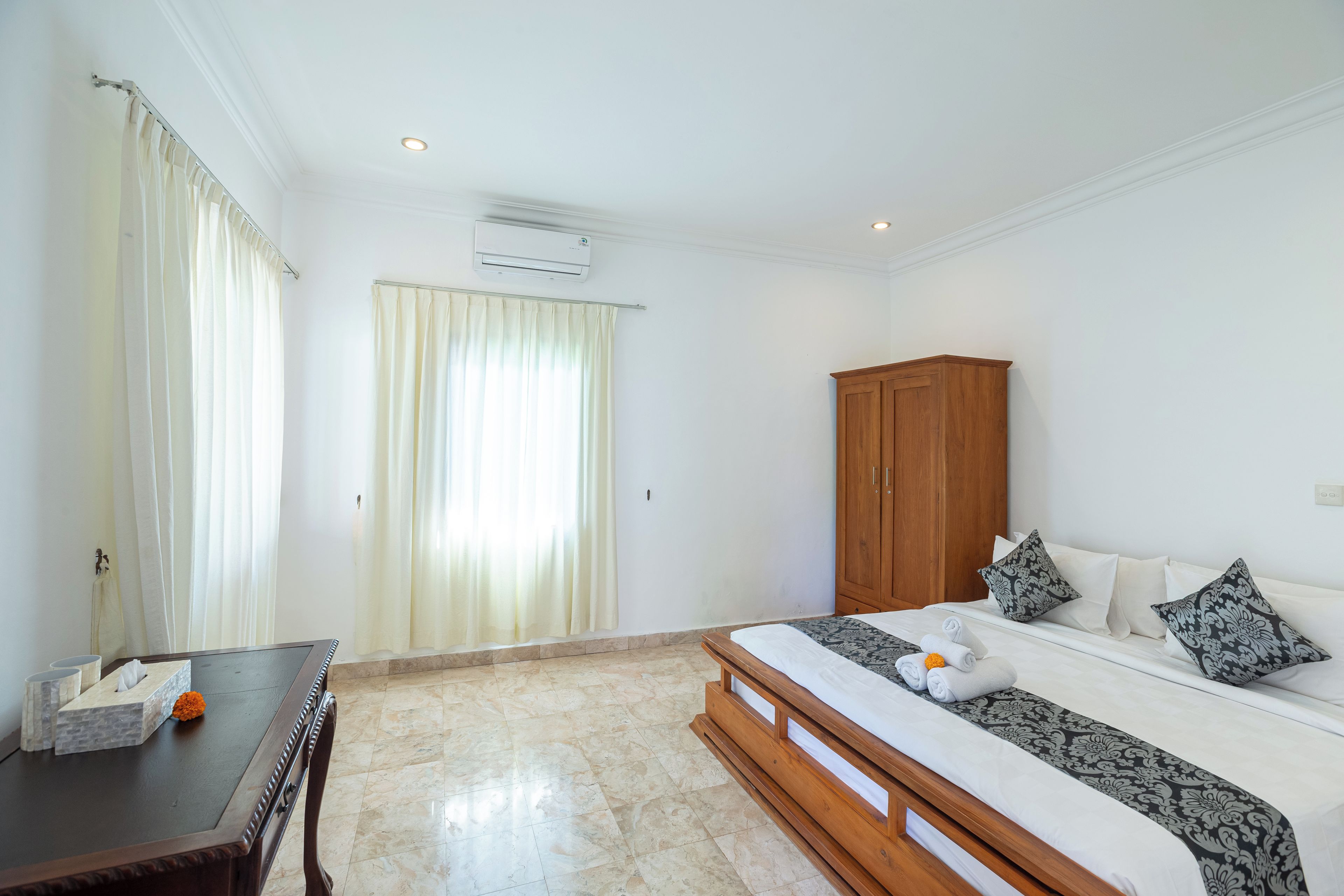 A hotel room with a bed, a wooden wardrobe, a desk with a tissue box and a flower, and a window with curtains.