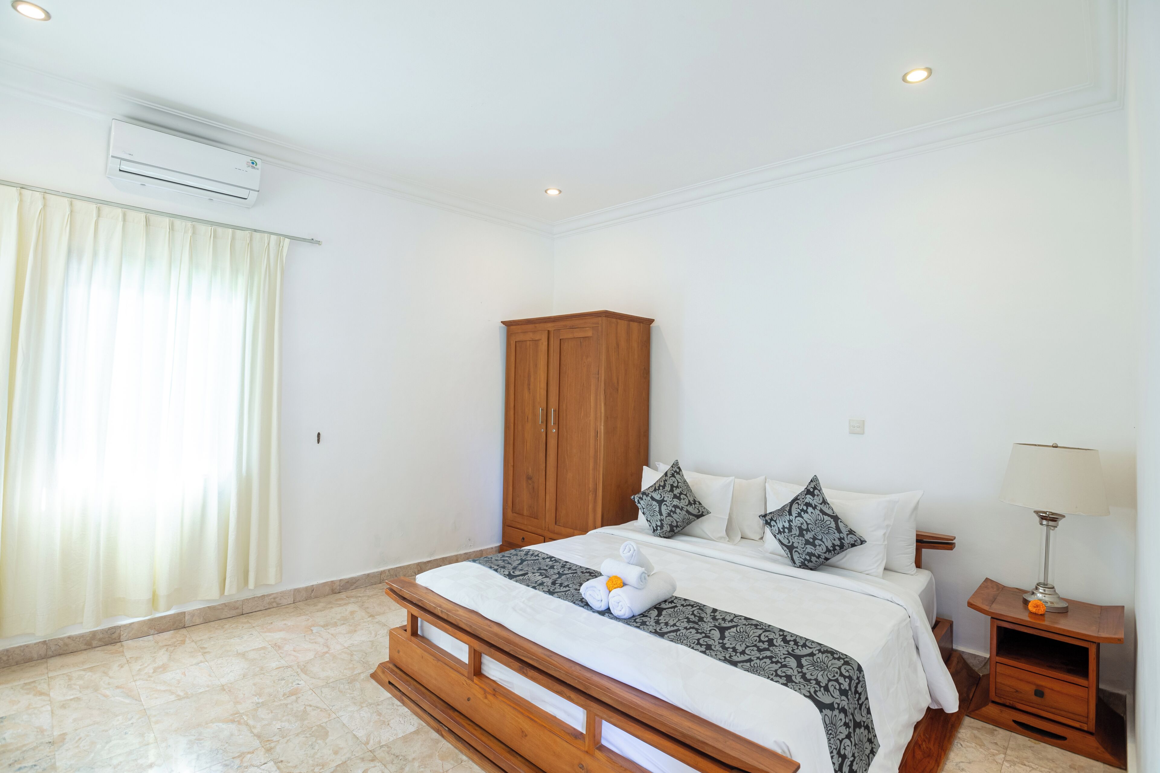 A bedroom with a bed, a wooden wardrobe, a nightstand with a lamp, and a window with curtains.