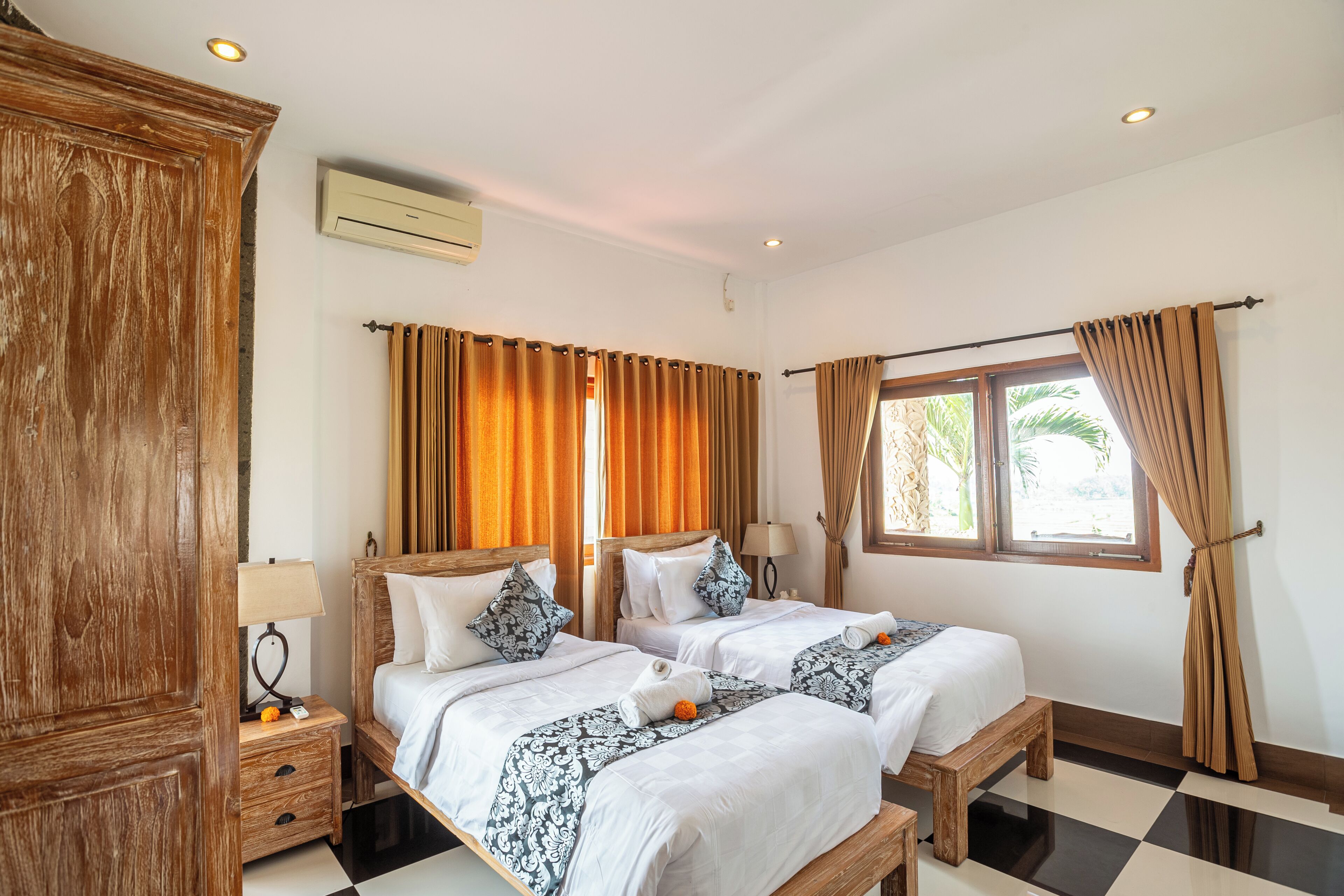 A hotel room with two beds, a wooden wardrobe, a window with curtains, and a ceiling fan.