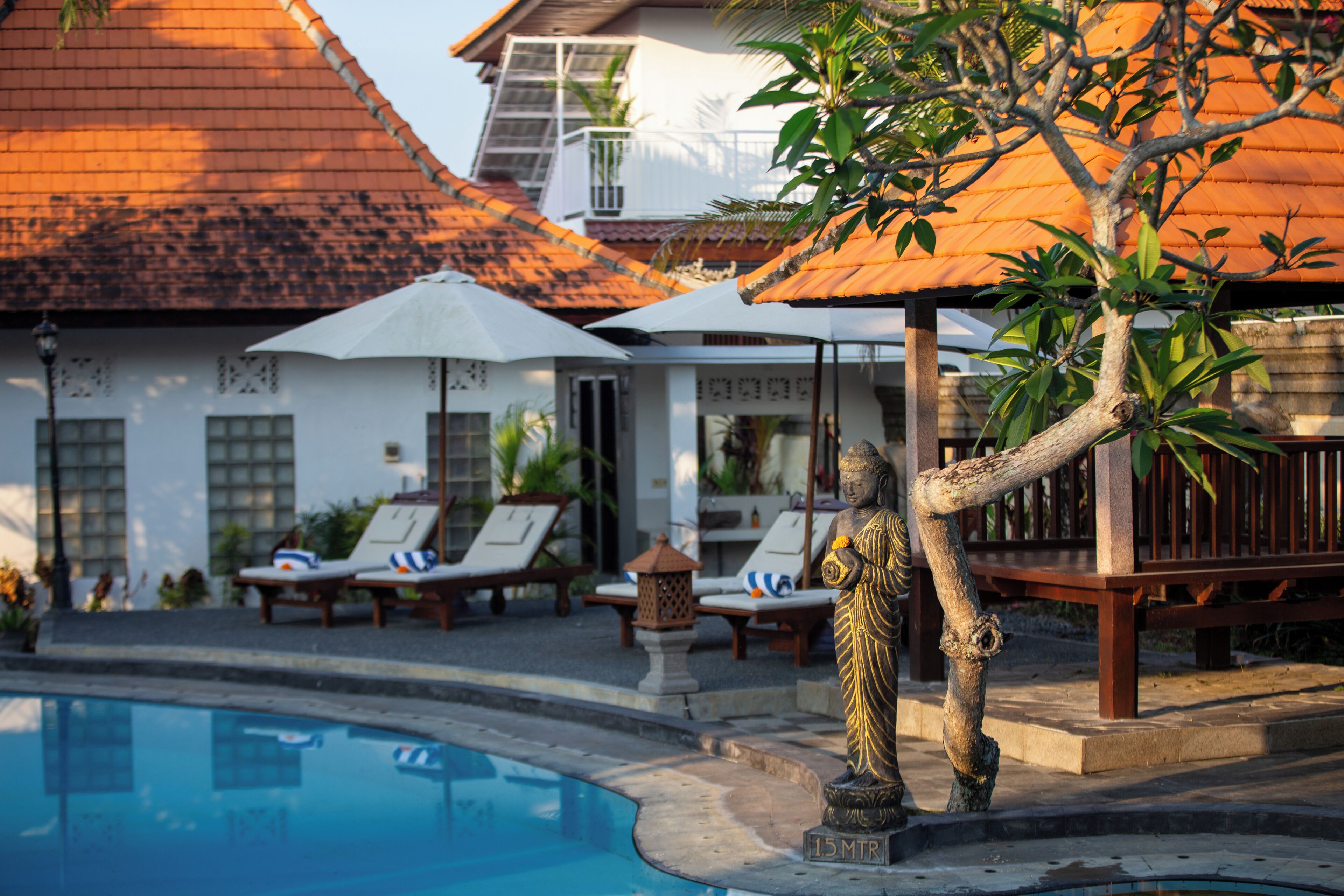 A pool area with lounge chairs and umbrellas, a Buddha statue, and a building with a tiled roof.