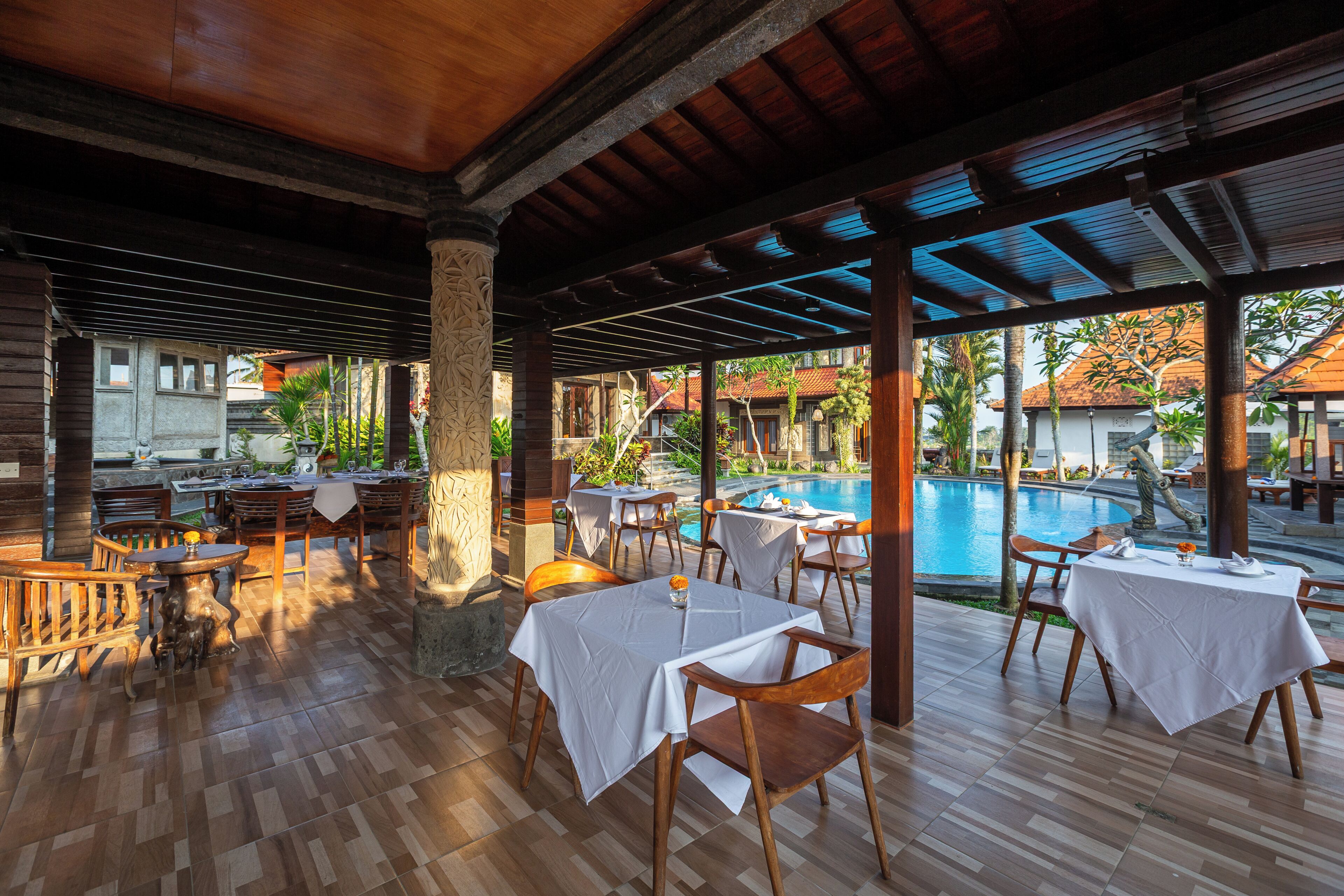 An outdoor dining area with wooden tables and chairs, a pool, and a view of buildings in the background.