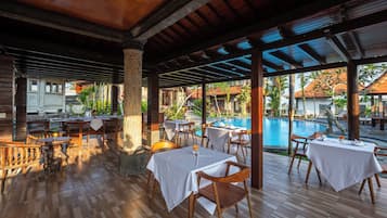 An outdoor dining area with wooden tables and chairs, a pool, and a view of buildings in the background.