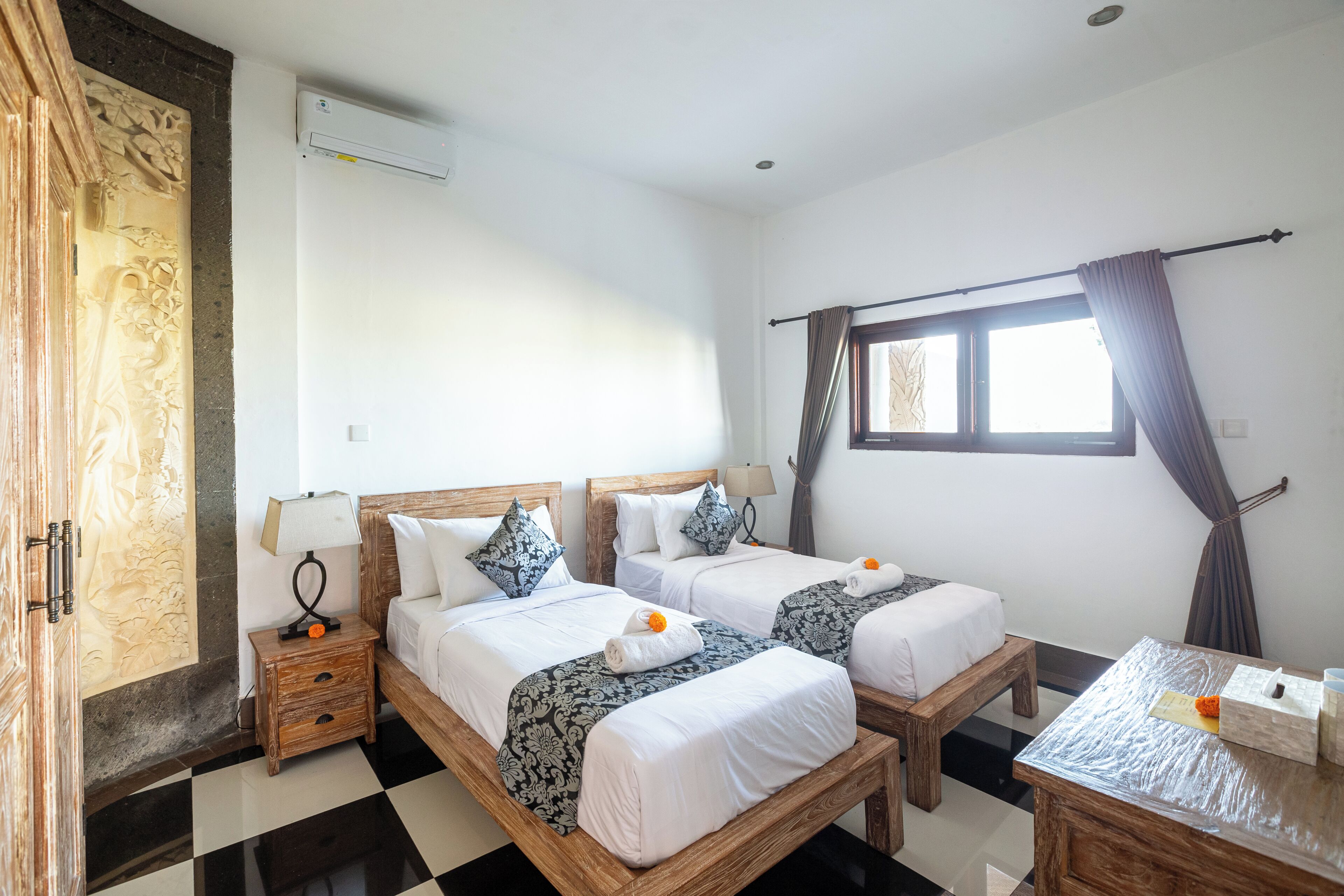 A hotel room with two beds, a wooden wardrobe, a small table, and a window with curtains.