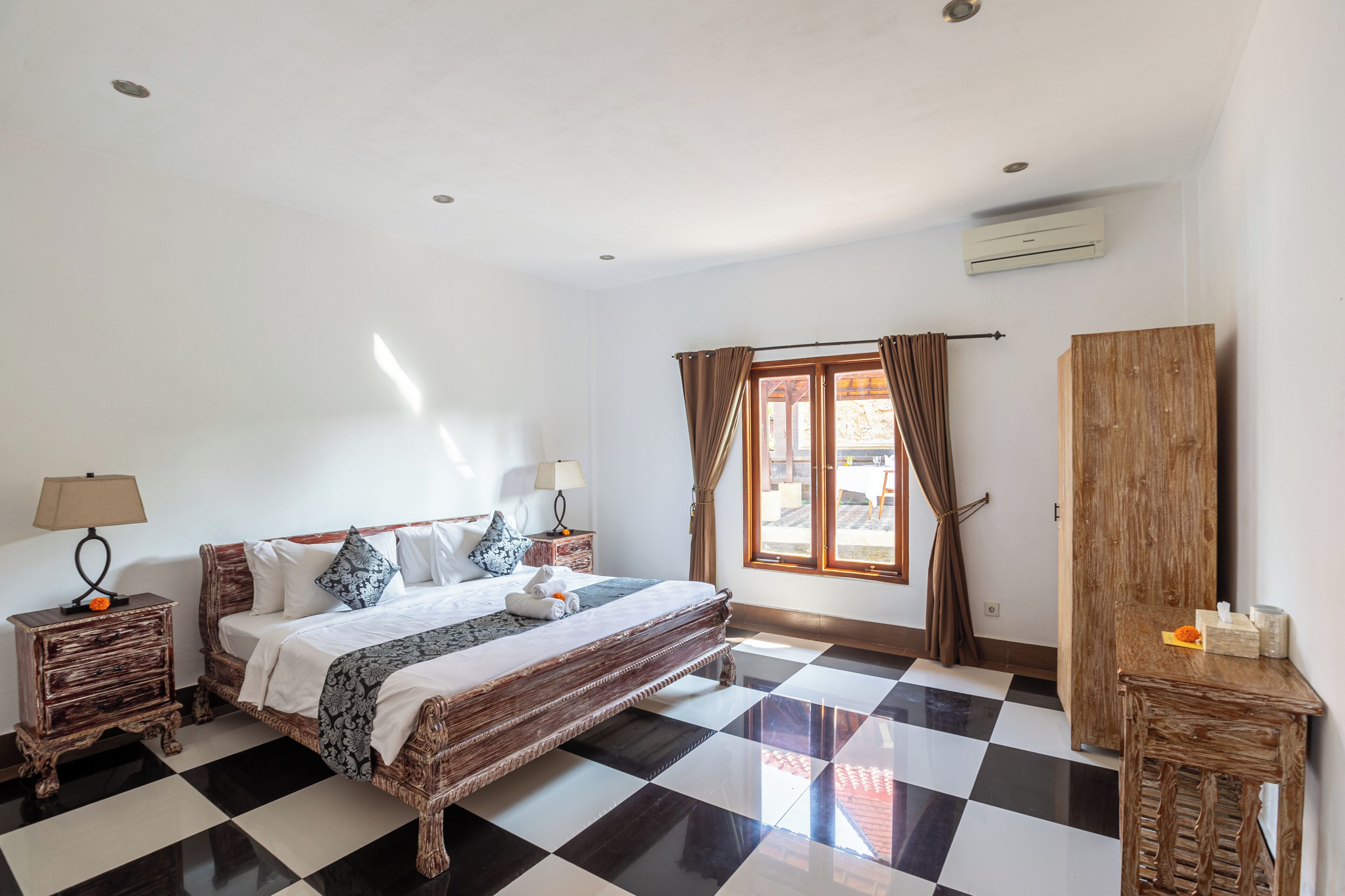 Ein Hotelzimmer mit Schachbrettmuster-Boden, einem Bett mit gemusterten Betttextilien, einem Holzschrank und einem Fenster mit Vorhängen.