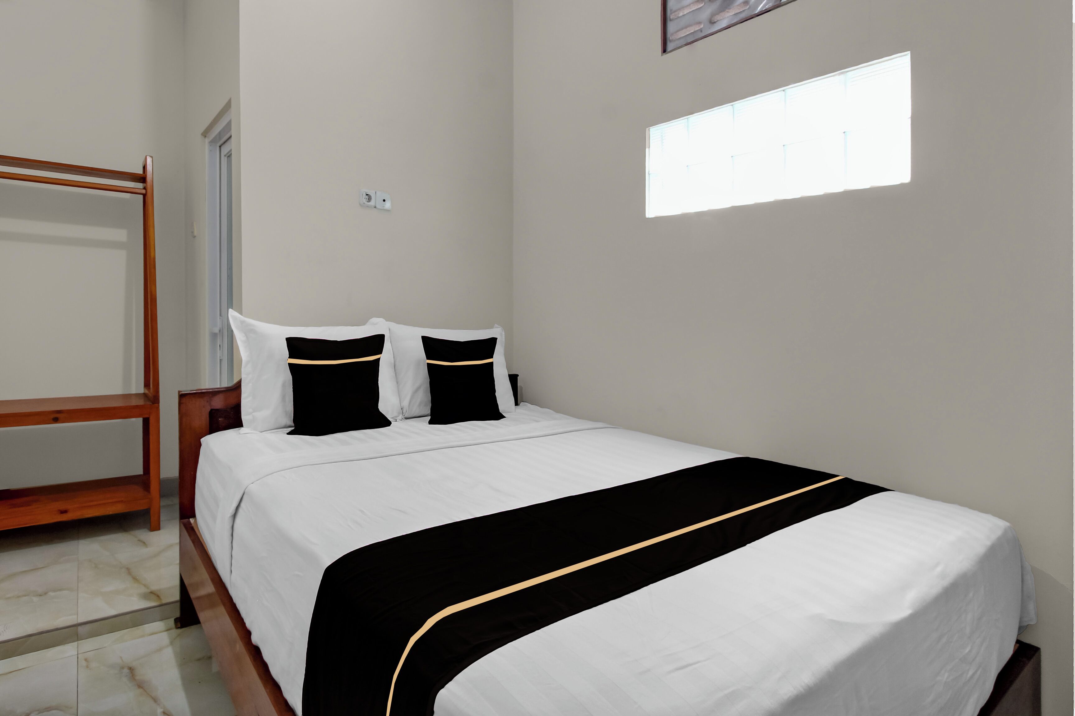 Una cama bien hecha con ropa de cama blanca y detalles negros, una mesita de noche de madera y una ventana que deja entrar luz natural.