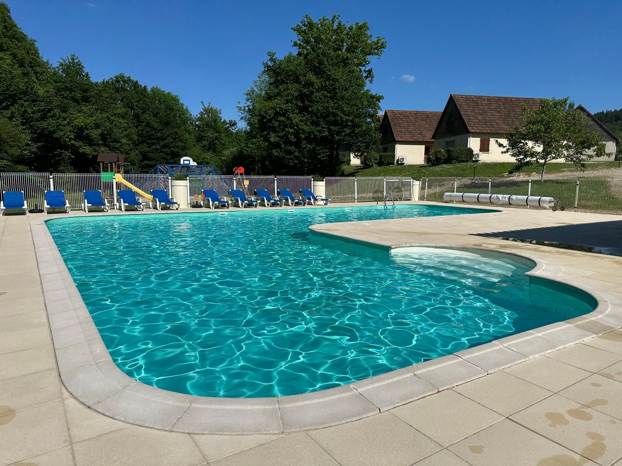 A swimming pool with clear blue water, surrounded by lounge chairs and a fenced area.
