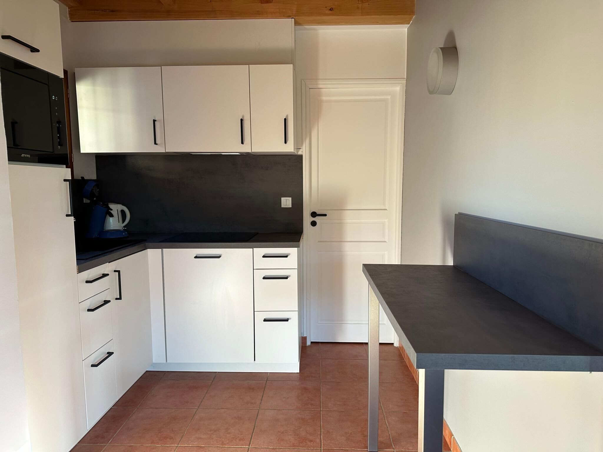 Een kleine keuken met witte kastjes, een zwart aanrecht en een grijze bank.