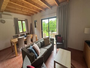 A cozy living room with a sofa, dining table, and chairs, a large window with curtains, and a wooden ceiling. - Beautiful house in a holiday park with a pool (Lacapelle Marival)