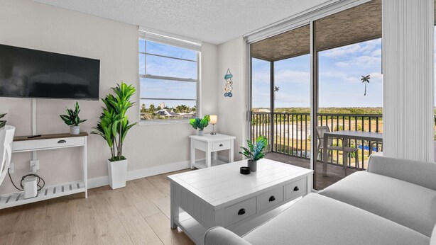 Ein modernes Wohnzimmer mit Flachbildfernseher, weißen Möbeln, Blumentöpfen und einem Balkon mit Blick auf den Strand.